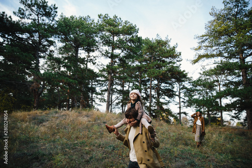 Young father and kid girl sitting on his neck, mother in a beret and a coat are having fun in the autumn forest.