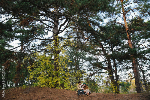 Young father, mother and little toddler daughter girl in a beret and a coat sitting on the ground in the autumn forest. photo