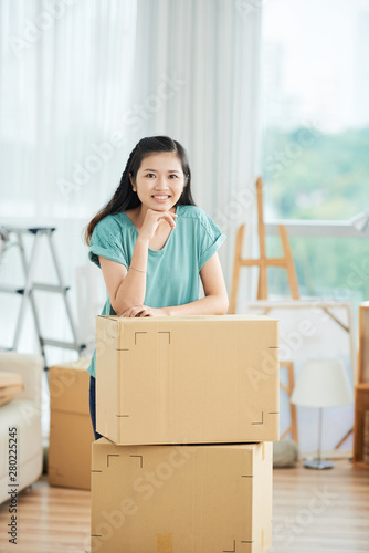 Portrait of Asian young woman standing near the packed cardboard boxes and smiling at camera at home