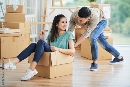 Happy Asian woman sitting in big cardboard box while her boyfriend pushing the box they having fun during their moving