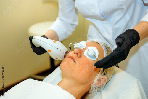 Doctor in medical gloves applies the patient special gel. Anti acne phototherapy with professional equipment. Young woman during procedure of rejuvenation. Facial skin treatment in a cosmetic clinic.