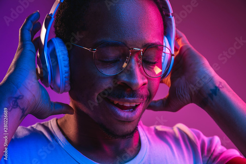 Young african-american man's listening to music in headphones in neon light on gradient background. Male portrait. Concept of human emotions, facial expression, summer holidays or weekend, hobby,