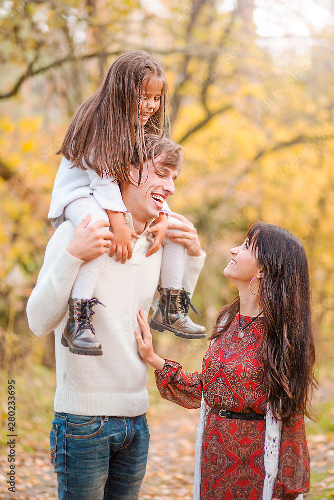 Obraz premium Mom, dad and daughter walk through the autumn forest. Daughter sits on father's shoulders