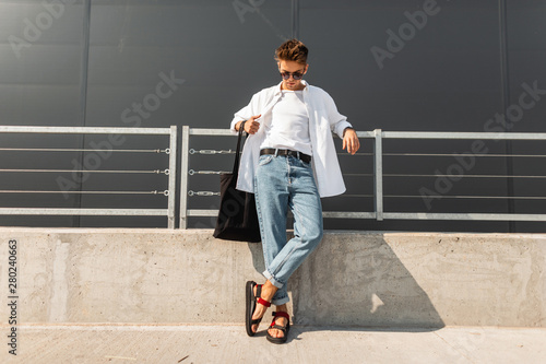 Modern young man hipster in fashionable summer white clothes in sunglasses in stylish red leather sandals with a bag stands on a sunny day near a gray building. Nice guy. Trendy menswear. Street style photo