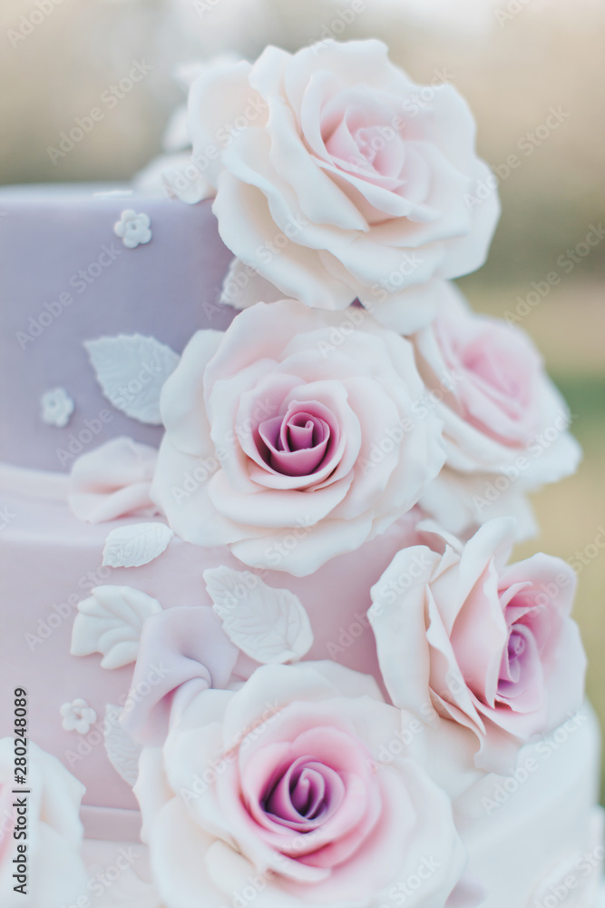 Close-up of parts of a three tiered wedding cake in pastel colors decorated with realistic pink roses on a blurred background of the garden, selective focus