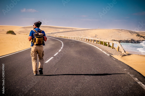 Man walking in the middle of a long way road with desert on both side and ocean beach - concept of travel and alternative lifestyle with backpack discovering the world with wanderlust