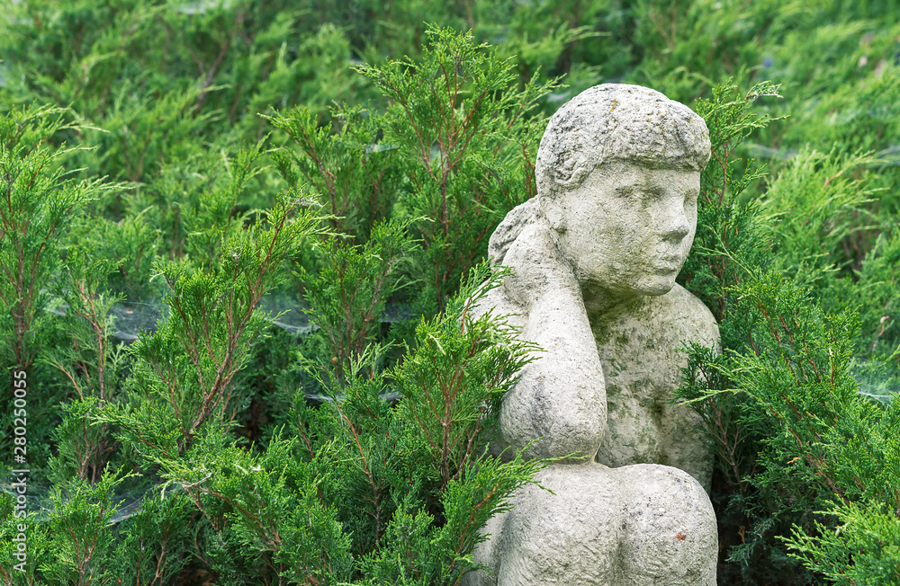 An ancient stone statue of a sitting girl among the green shrubs.