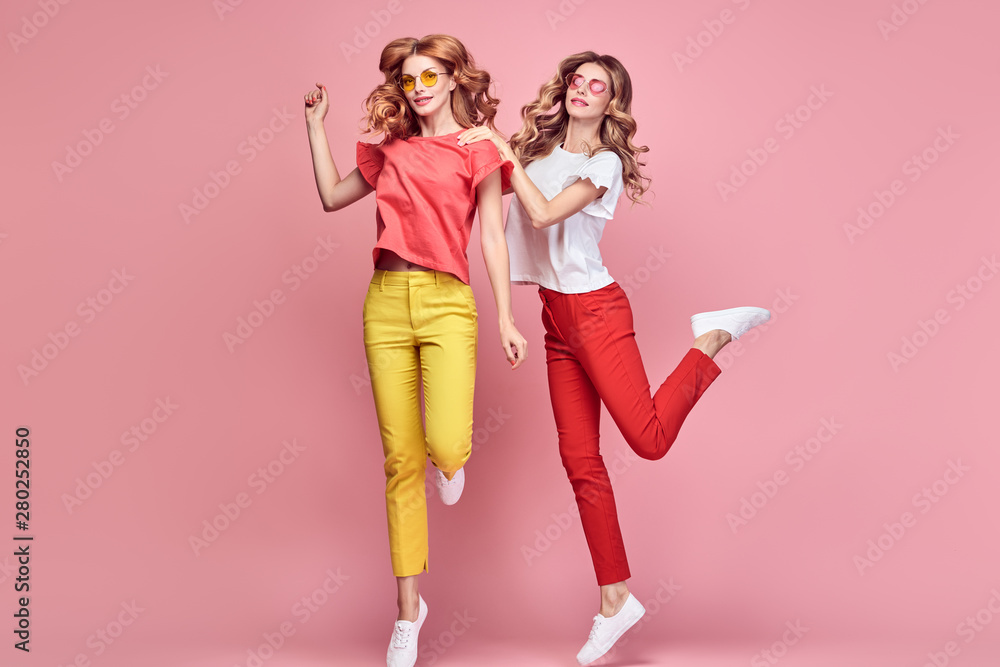 Two fashionable girl jump Smiling in colorful outfit on pink. Beautiful easy-going woman in red yellow pants, Stylish curly hair having fun. Joyful funny slim sisters friends, happy fashion concept