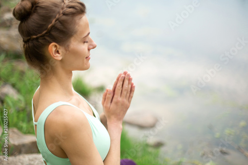 Thin brunette girl plays sports and performs beautiful and sophisticated yoga poses in a summer park. Green forest and the river on the background. Woman doing exercises on the yoga mat. Lotus pose