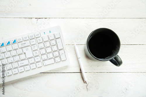 Black cup with coffee and computer keyboard on a white table. White wooden background. Board of boards