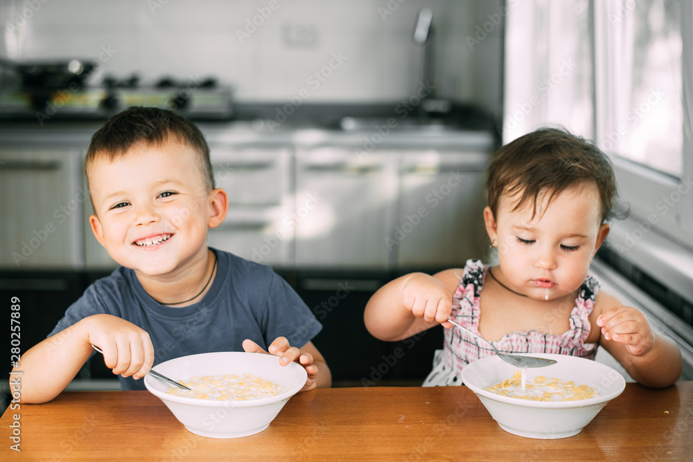 Brother and sister eat cornflakes with milk in the kitchen during the day very fun and cute, dirty and funny