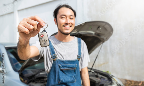 Portrait Of Asian young man auto mechanic In garage holding car key. Mechanic boy handling key to the customer. Repair of machines, fault diagnosis, specialist, technical maintenance service concept