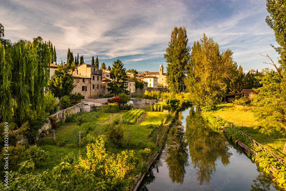The Chiasco river that crosses the ancient medieval village of Bevagna. Perugia, Umbria, Italy. Trees, vegetation, cultivated gardens. The blue sky at sunset, in the summer.