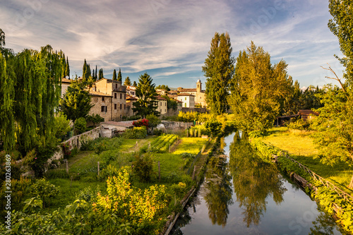Fototapeta Naklejka Na Ścianę i Meble -  The Chiasco river that crosses the ancient medieval village of Bevagna. Perugia, Umbria, Italy. Trees, vegetation, cultivated gardens. The blue sky at sunset, in the summer.