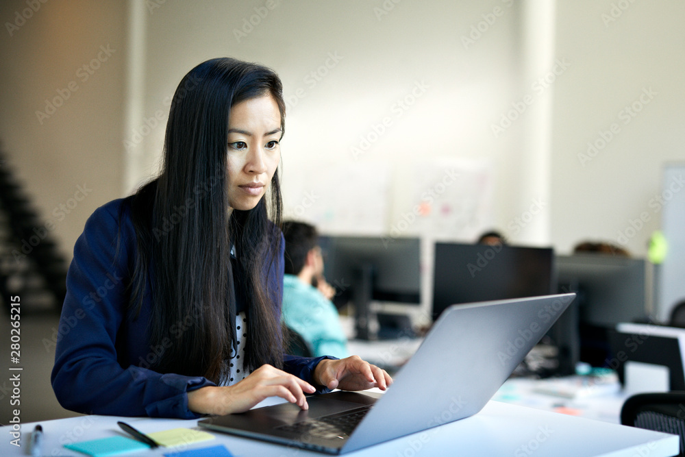 Confident businesswoman using laptop computer on desk while standing against wall in office Confident businesswoman using laptop computer on desk while standing against wall in office
