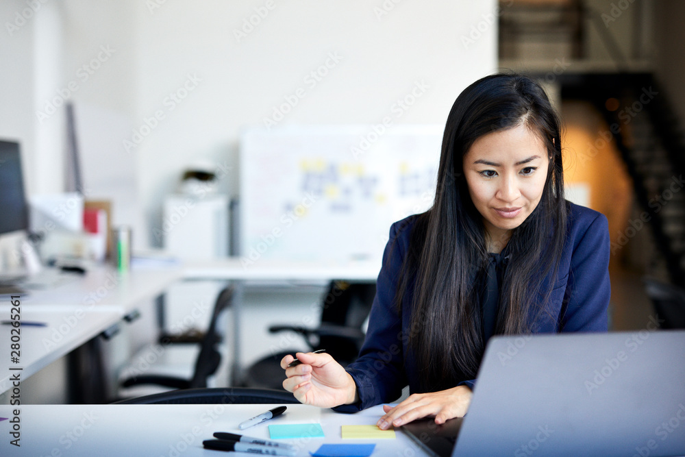 © Cavan Images - Confident businesswoman writing on adhesive notes while using laptop computer against wall in office