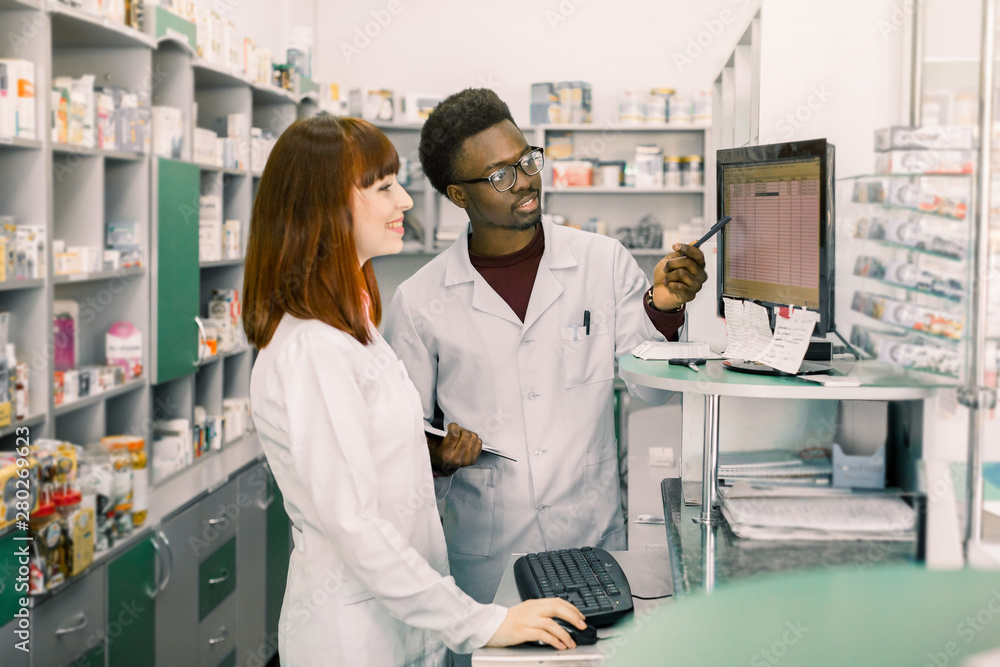 Obraz premium Team of two young apothecaries in pharmacy standing in front of shelves with drugs and working on computer making order of medicines