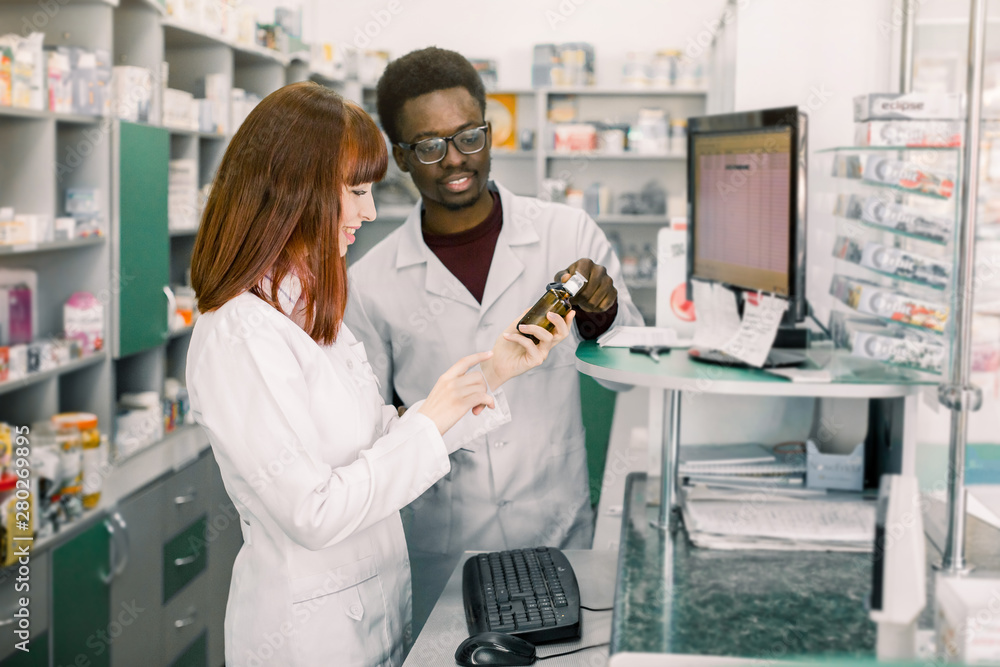 Obraz premium Young Caucasian female pharmacist talking with her African man colleague about the new medicine standing near the counter and PC, in the pharmacy
