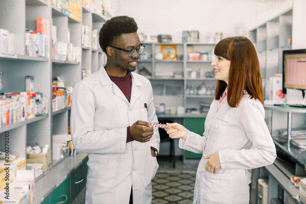 Confident Male And Female Pharmacists In Pharmacy. African man and Caucasian woman holding blister of pills while standing in modern pharmacy