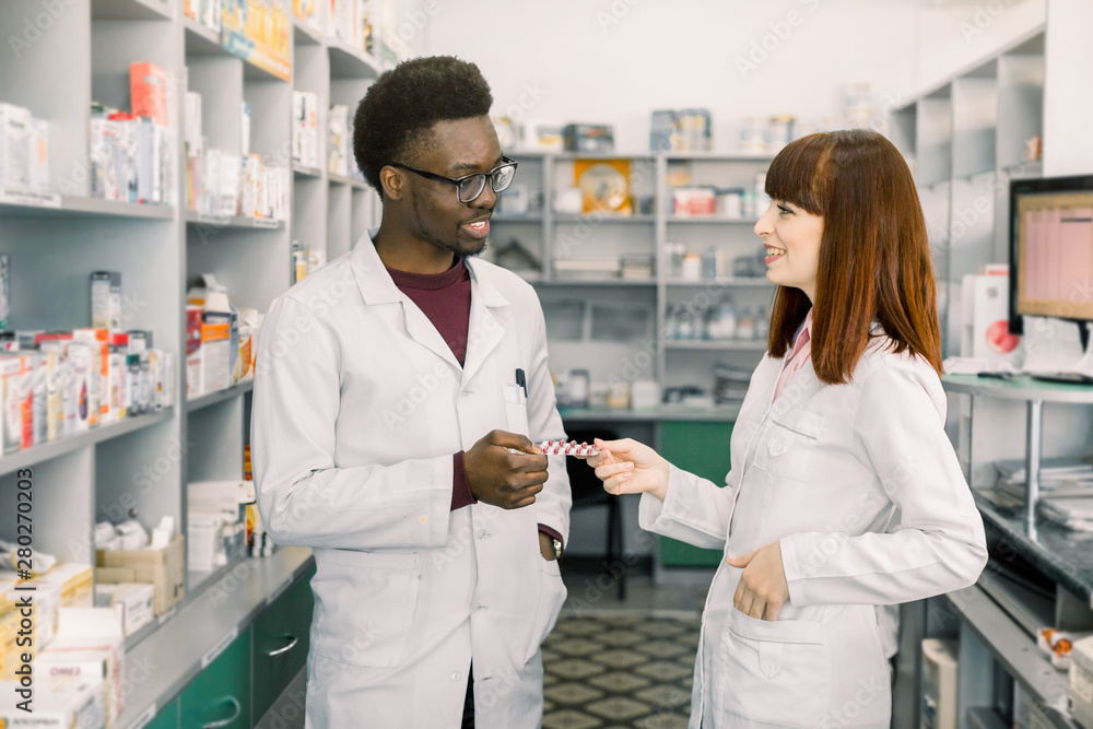 Obraz premium Confident Male And Female Pharmacists In Pharmacy. African man and Caucasian woman holding blister of pills while standing in modern pharmacy