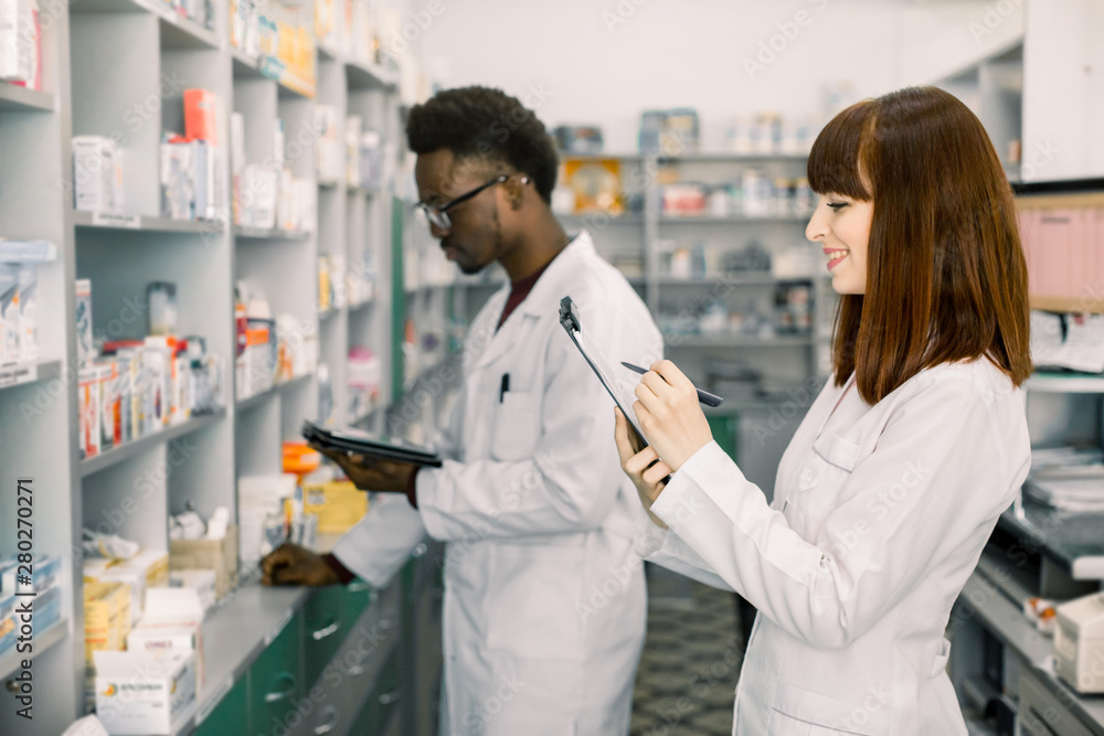 Confident Male And Female Pharmacists In Pharmacy. African American male pharmacist working on digital tablet and Caucasian woman making notes on clipboard during inventory in pharmacy