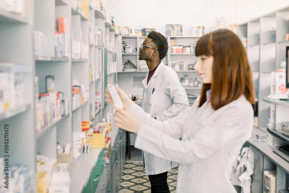 Two young multiethnical Pharmacists Working at modern pharmacy. Female Pharmacist Holding package with Pills, male pharmacist standing on the background. Pharmacists Wearing Medical Uniform.