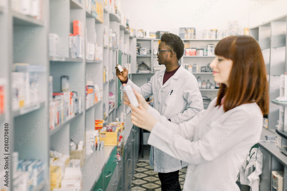 Two Pharmacists Working. Caucasian Woman and African Man Wearing Special Medical Uniform. Woman Showing package with Pills. Man looking for medicines on Background.