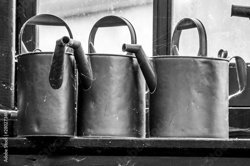 Three wattering cans on the shelve by the window in black and white photo