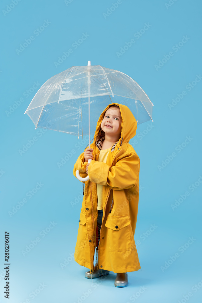 Happy funny child with transparent umbrella posing on blue studio  background. Girl is wearing yellow raincoat and rubber boots. Holds a  vintage travel suitcase Stock Photo | Adobe Stock
