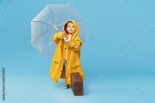 Happy funny child with transparent umbrella posing on blue studio background. Girl is wearing yellow raincoat and rubber boots. Holds a vintage travel suitcase