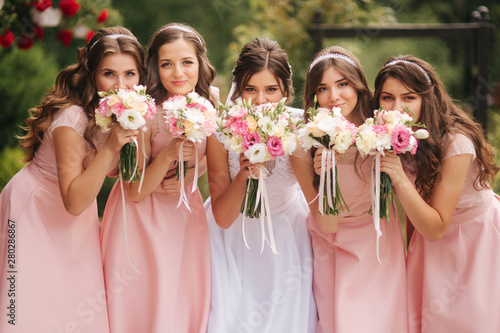 Happy bride with bridesmaid hold bouquets and have fun outside. Beautiful bridesmaid in same dresses stand by the charming bride in long wedding dress