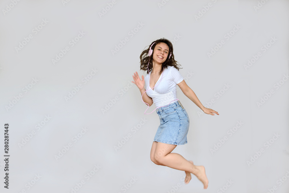 close up photo of a happy brunette with pink headphones in white blouse and denim skirt jumping in the air