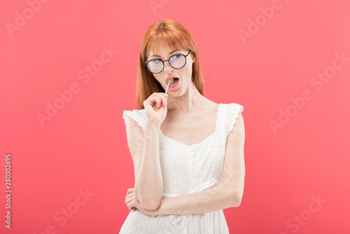 pensive girl in glasses eating lollipop and looking away isolated on pink