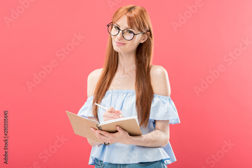 attractive redhead woman in glasses holding pencil and textbook isolated on pink