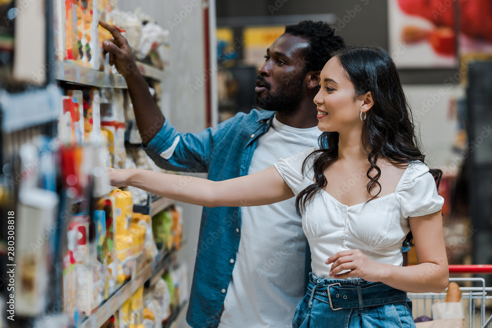 selective focus of handsome african american pointing with finger near attractive asian woman looking at shelves with groceries