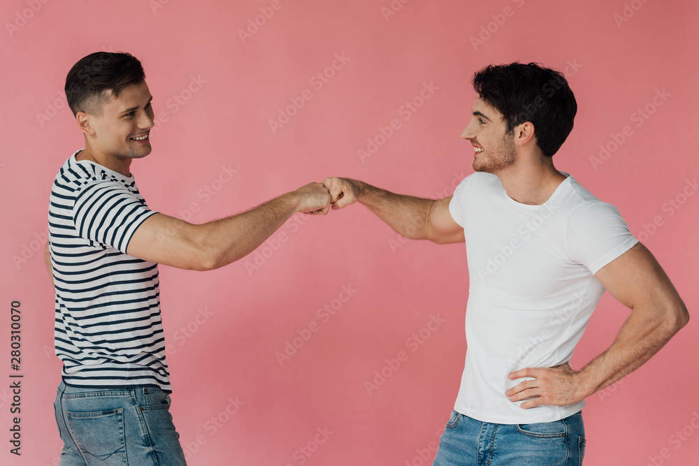 two smiling friends in t-shirts touching fists and looking at each other isolated on pink two smiling friends in t-shirts touching fists and looking at each other isolated on pink
