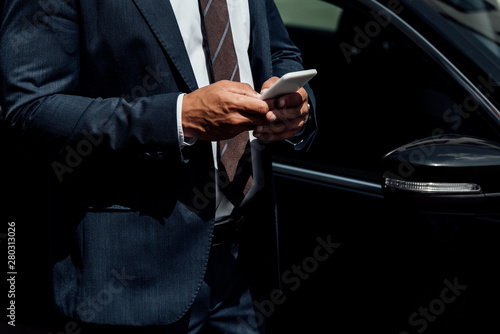 cropped view of african american businessman in suit using smartphone at sunny day near car