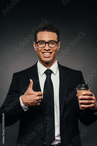 handsome smiling african american businessman in glasses and suit holding coffee to go and showing thumb up on dark background