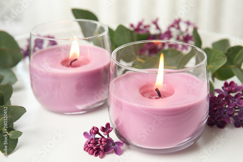 Burning candles in glass holders and flowers with leaves on white table