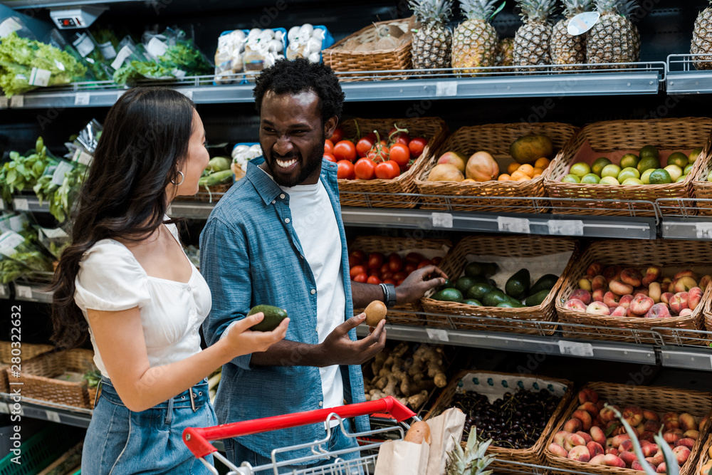 happy asian girl holding avocado near shopping cart and african american man with kiwi fruit in supermarket