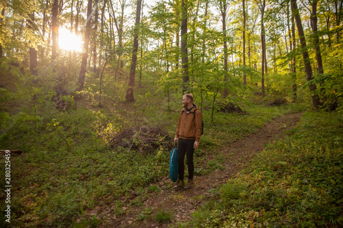 one young man, walking in forest, while some camping equipment. smiling and looking at sunlight.