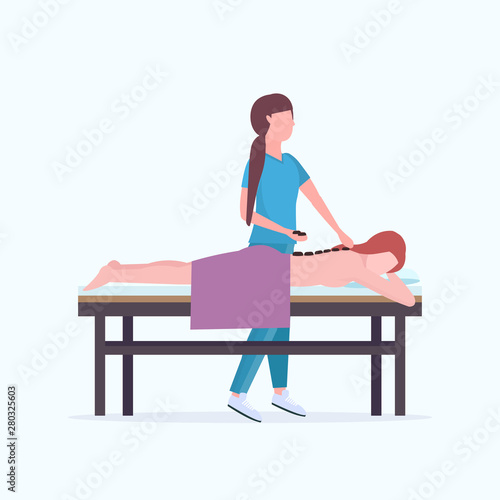 young girl having hot stone back massage masseuse in uniform massaging patient body woman relaxing lying on bed luxury spa salon treatments concept full length
