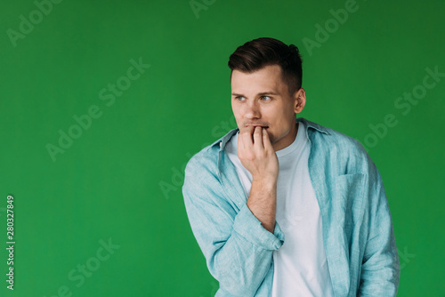 worried young man in shirt biting fingers and looking away isolated on green