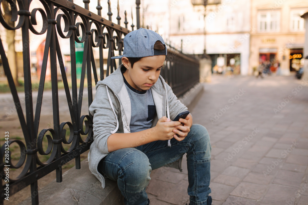 Young boy play online game at smartphone outside. Boy sitting in centre of the city. Boy play games while his mom going to the shop Young boy play online game at smartphone outside. Boy sitting in centre of the city. Boy play games while his mom going to the shop