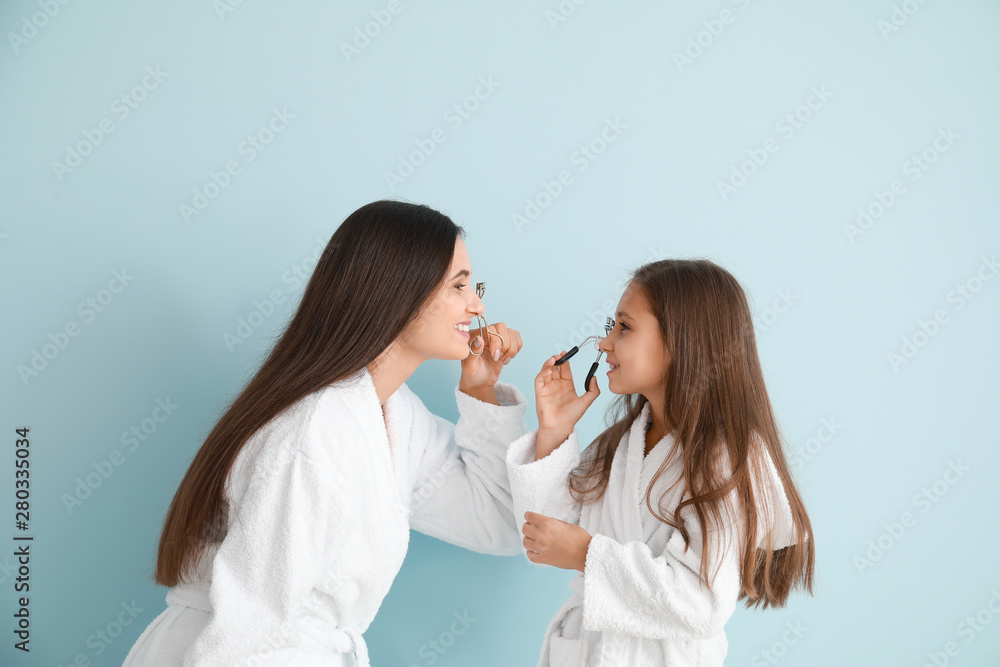 Mother and her little daughter in bathrobes using eyelash curlers on color background