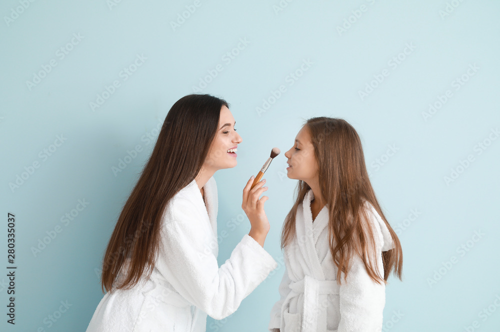 Mother and her little daughter in bathrobes applying makeup against color background Mother and her little daughter in bathrobes applying makeup against color background
