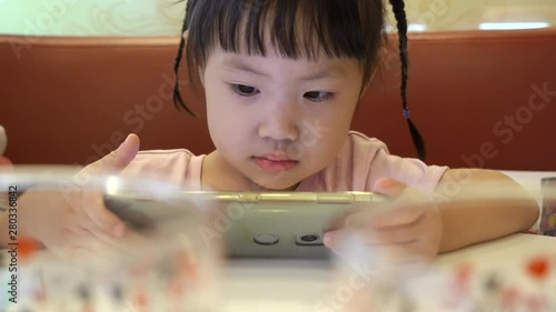 Asian baby girl watch cartoons in smartphone at restaurant in shopping mall. Handheld shot 4K