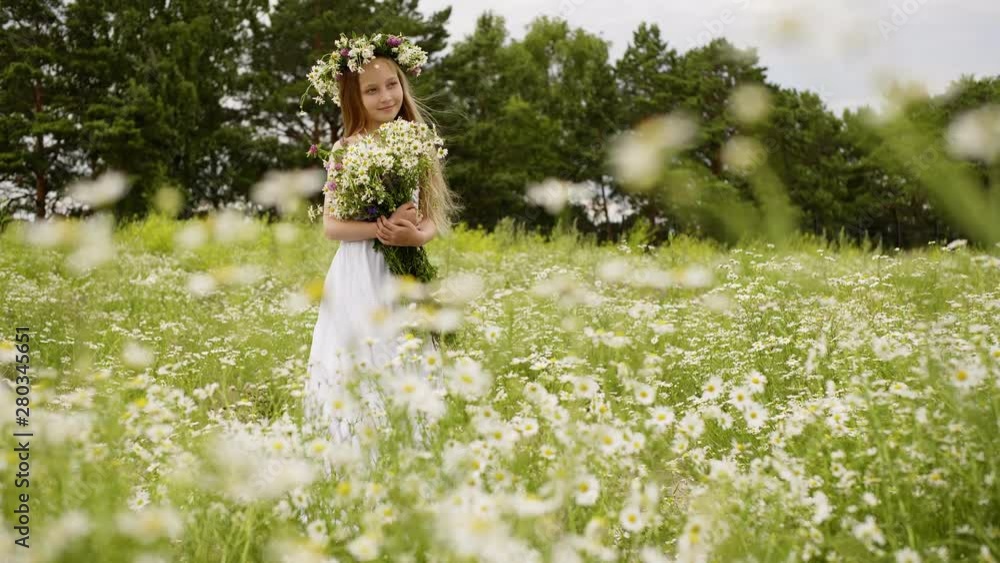 Smiling young girl in wreath with flowers bouquet standing on flowering field. Beautiful girl posing front camera on blooming meadow at summer windy day Smiling young girl in wreath with flowers bouquet standing on flowering field. Beautiful girl posing front camera on blooming meadow at summer windy day