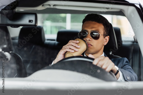 african american businessman in suit and sunglasses driving car and drinking coffee at sunny day