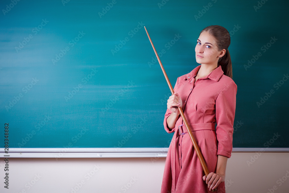 Young woman teacher stands at a blackboard with a pointer. Template for the school timetable and other information. Beautiful tacher shows on empty copy space on the school board. Education concept. Young woman teacher stands at a blackboard with a pointer. Template for the school timetable and other information. Beautiful tacher shows on empty copy space on the school board. Education concept.